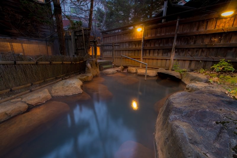 Outdoor Japanese hot spring bath surrounded by natural rocks and wooden fencing at dusk